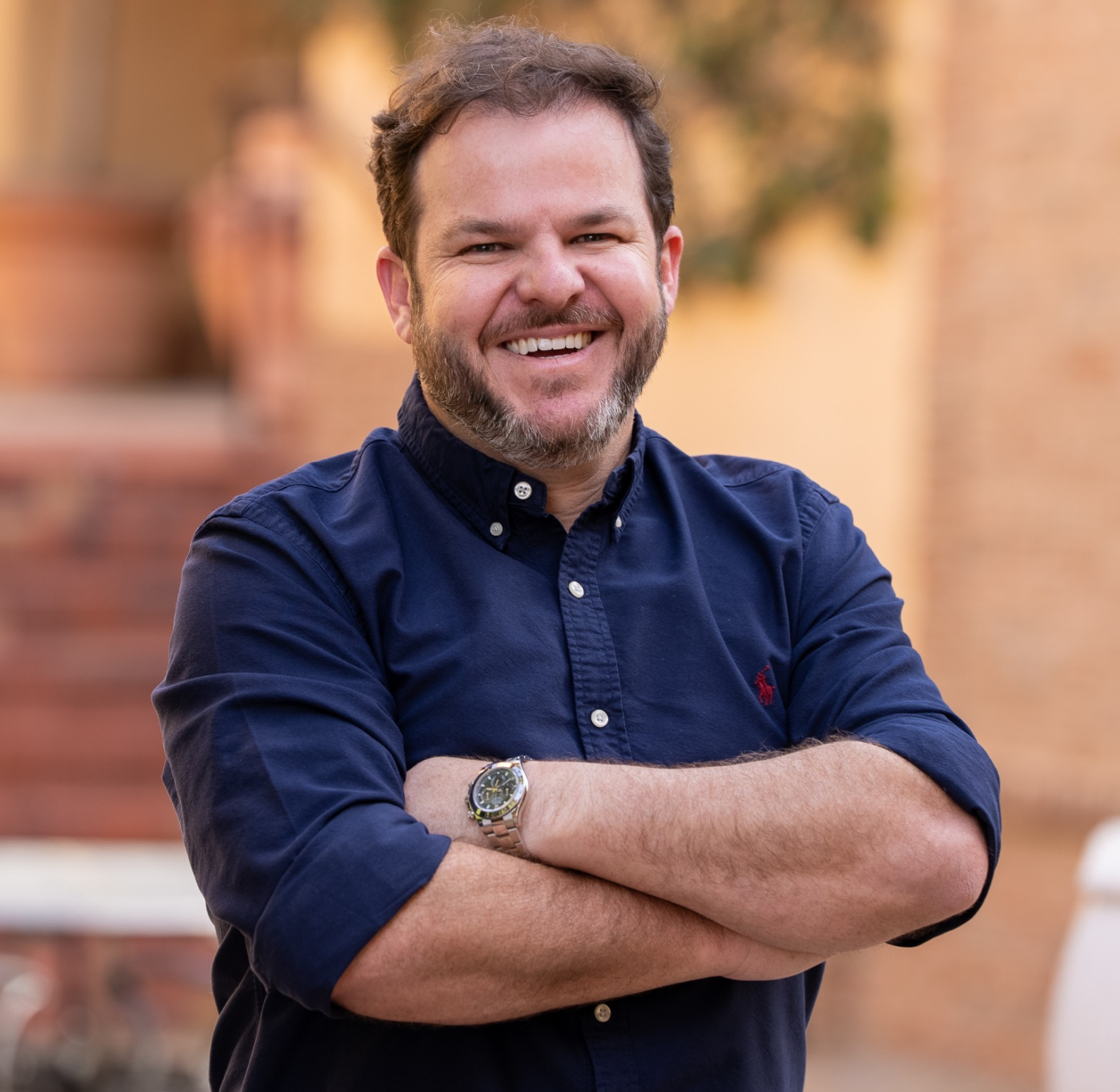 A man with a short beard, wearing a navy blue button-down shirt, smiles broadly with his arms crossed in front of a warm outdoor setting.