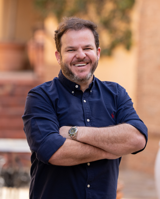 A man with a short beard, wearing a navy blue button-down shirt, smiles broadly with his arms crossed in front of a warm outdoor setting.